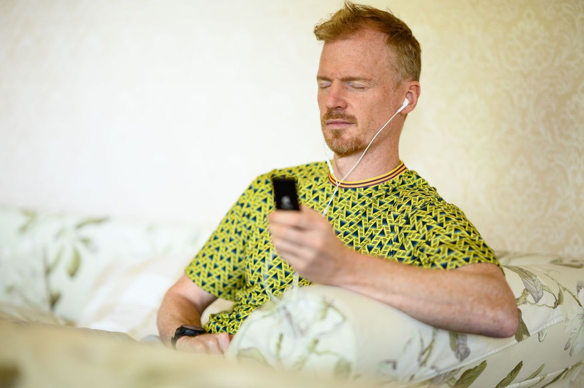 Man sat on a sofa holding a black iPod Nano (2006 8GB model) and his eyes are closed as he listens to the music